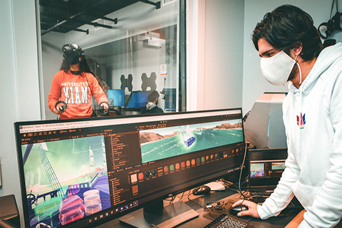 This is photo was taken on the University of Miami Coral Gables campus. A student is in the foreground editing a virtual reality app on a computer. Another student is in the background testing out the app. The student in the background is wearing virtual reality goggles.