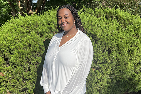 Nyanti poses in front of a bush, smiling at the camera. She is wearing a white shirt.